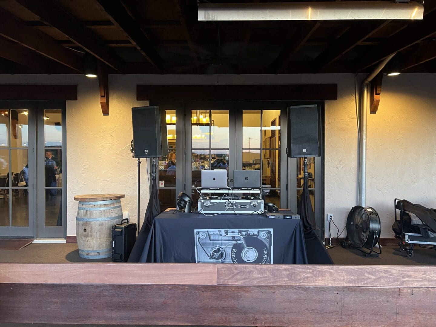 A DJ setup with two laptops and turntables on a table with black cloth between two large speakers on stands, positioned on a wooden deck. A wine barrel stands on the left side near glass doors.