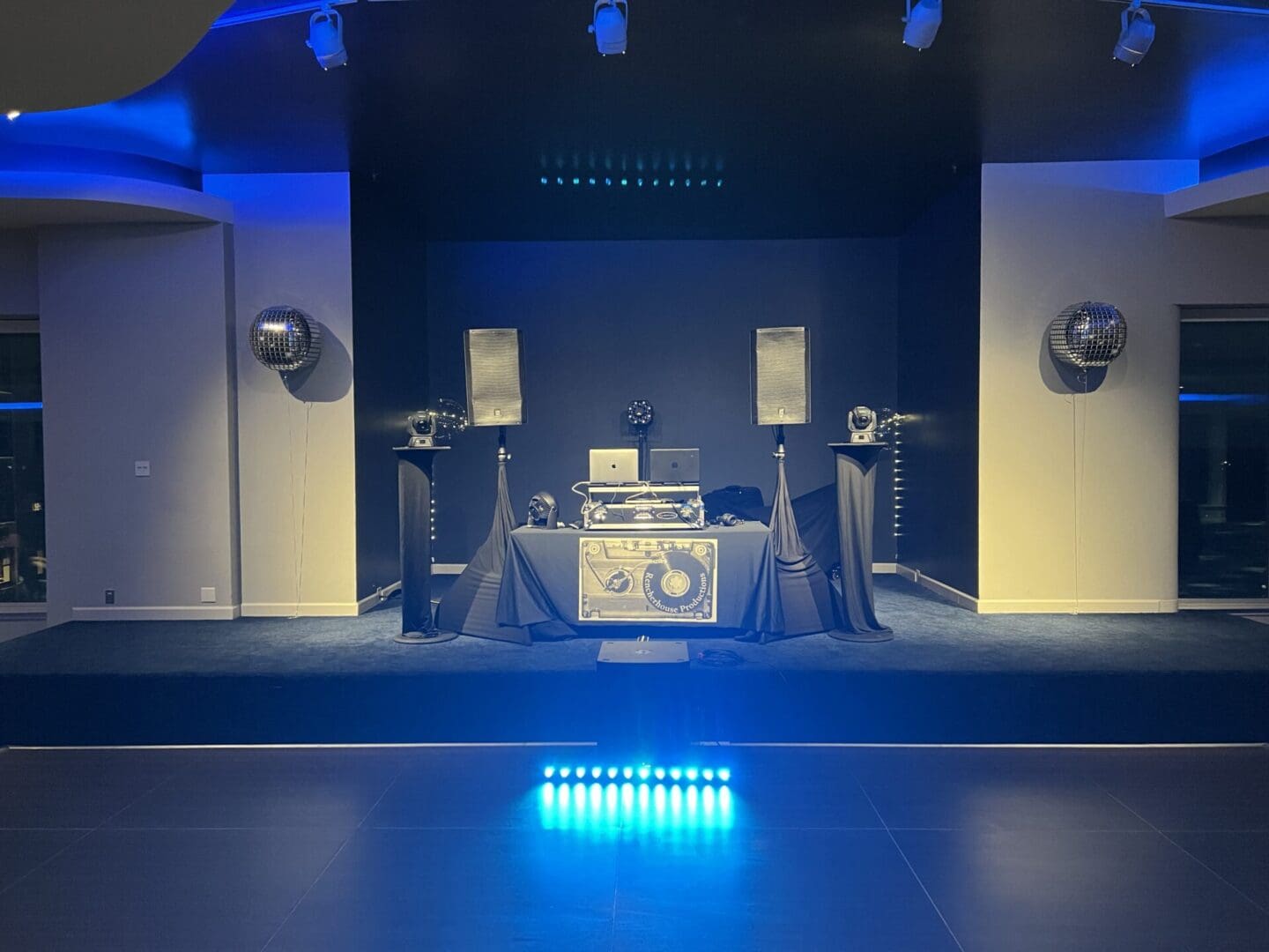 A DJ booth set up on a stage with speakers, turntables, and various equipment, flanked by disco balls and illuminated with blue lighting in a dimly lit event space.